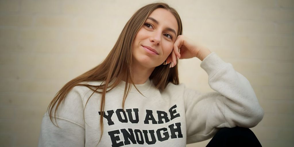 Young woman with long hair, wearing a "You Are Enough" sweatshirt, sits pensively with a gentle smile. The background is a soft, neutral color.