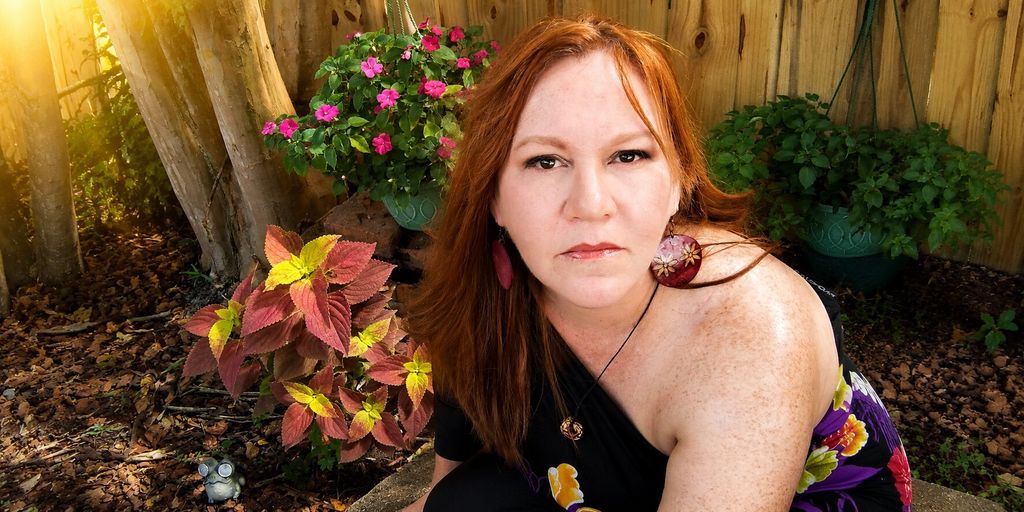 Red-haired woman seated in a sunlit garden surrounded by colorful plants and flowers, looking at the camera.