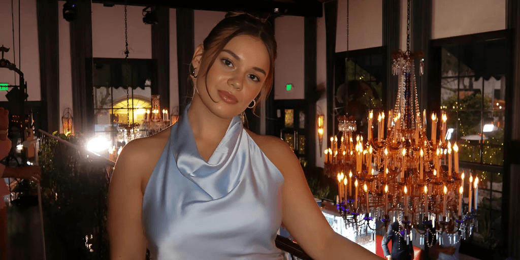 A woman in a satin blue dress smiles softly in front of chandeliers, exuding warmth and authenticity.