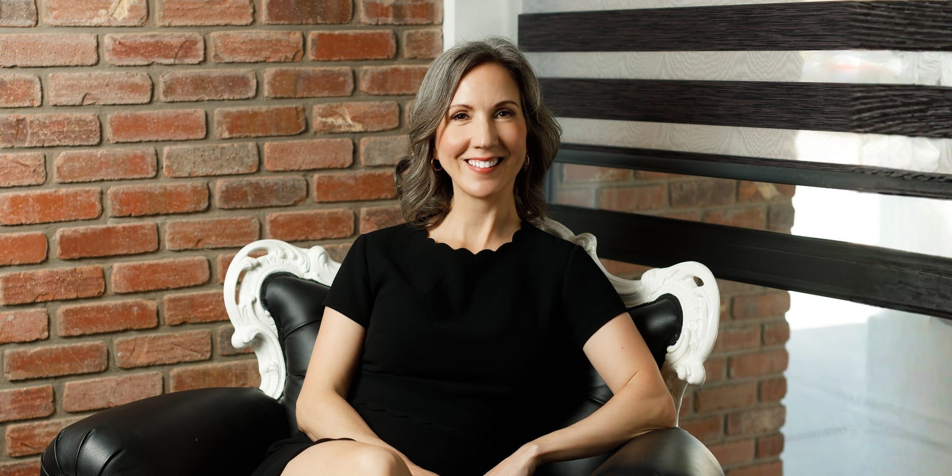 Smiling orthodontist seated in elegant office with brick wall background.