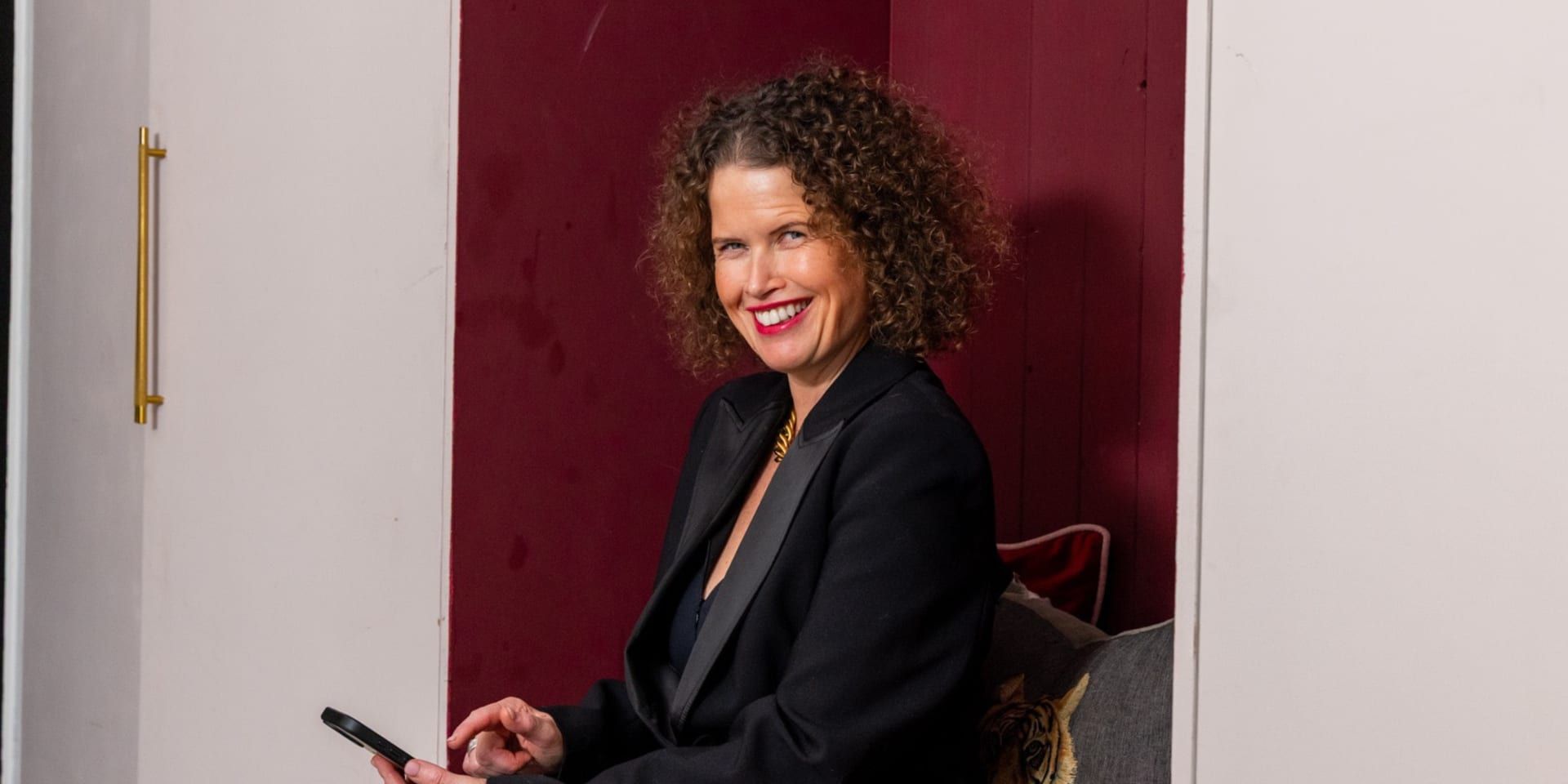 Dr Kate Armstrong smiling while seated with a smartphone, wearing a black blazer in a modern interior.