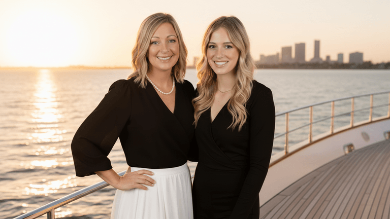 Two women in black outfits smiling on a yacht at sunset with city skyline in background.