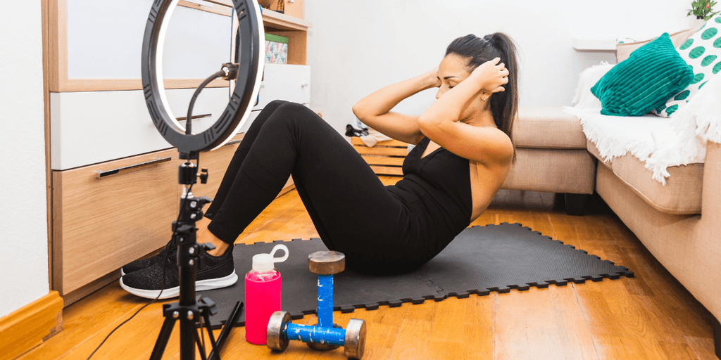 Woman exercising at home while filming a fitness workout with ring light, dumbbells, and sportswear.