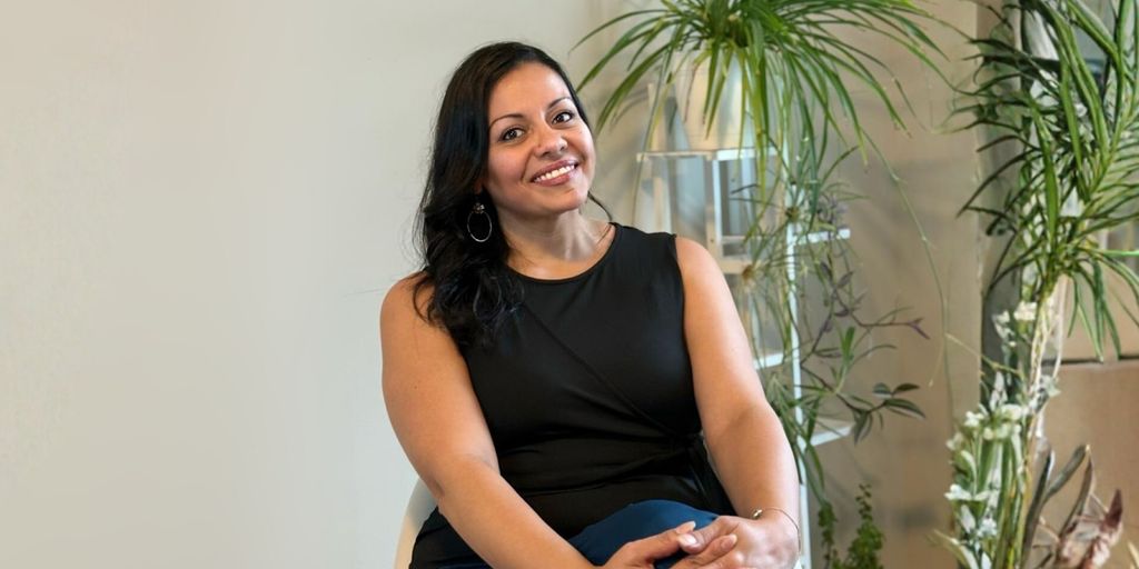Ellie smiling confidently, dressed in black, seated with plants in the background, embodying her transformational coaching journey.