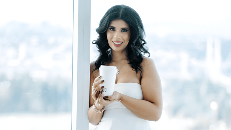 Priya Jhajj, founder of NeuroSomatics, standing confidently with a coffee cup in a bright, modern office, embodying a blend of science and holistic healing