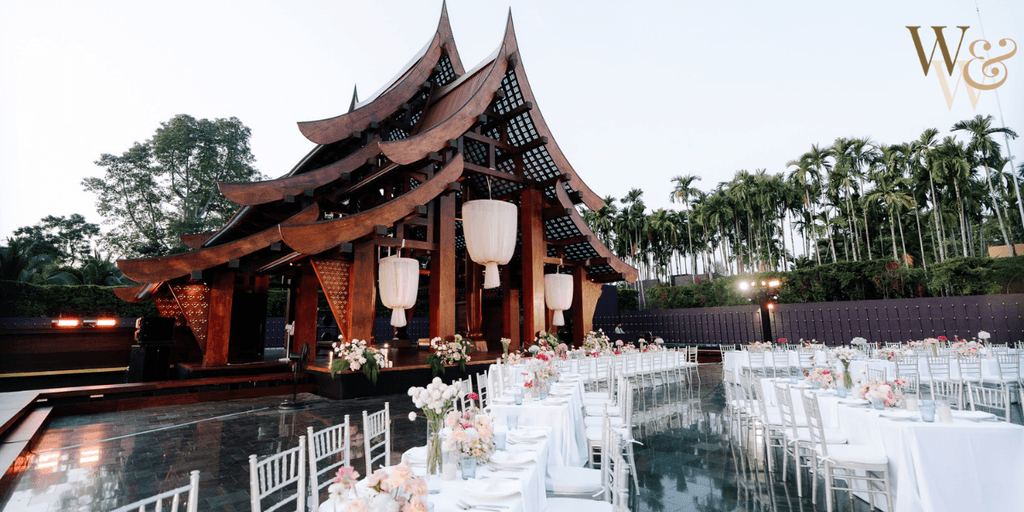 Outdoor wedding venue with a traditional wooden pavilion, long dining tables adorned with floral arrangements, and a scenic backdrop of palm trees under a clear sky.