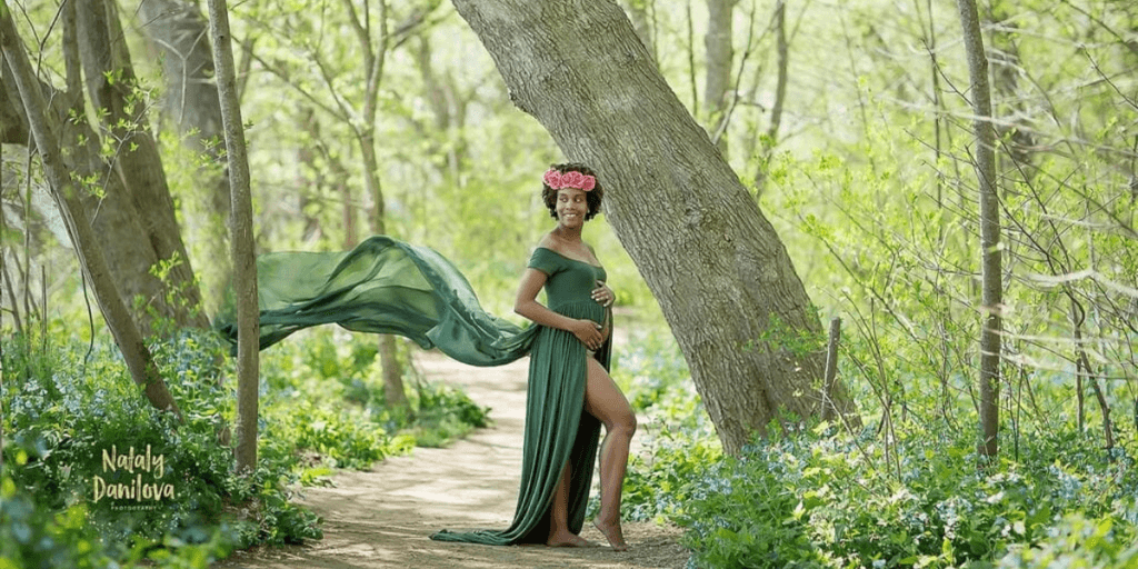 "Pregnant woman in a flowing green dress standing confidently in a nature-filled path with a flower crown, exuding elegance and grace during a maternity photo shoot."