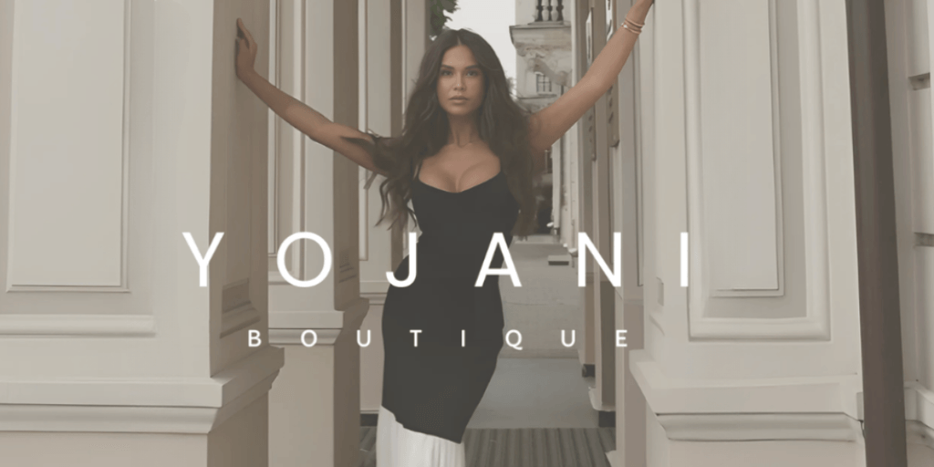 Fashion model posing confidently in a black dress, standing between elegant columns with the 'Yojani Boutique' logo displayed prominently, showcasing sophisticated and stylish clothing.