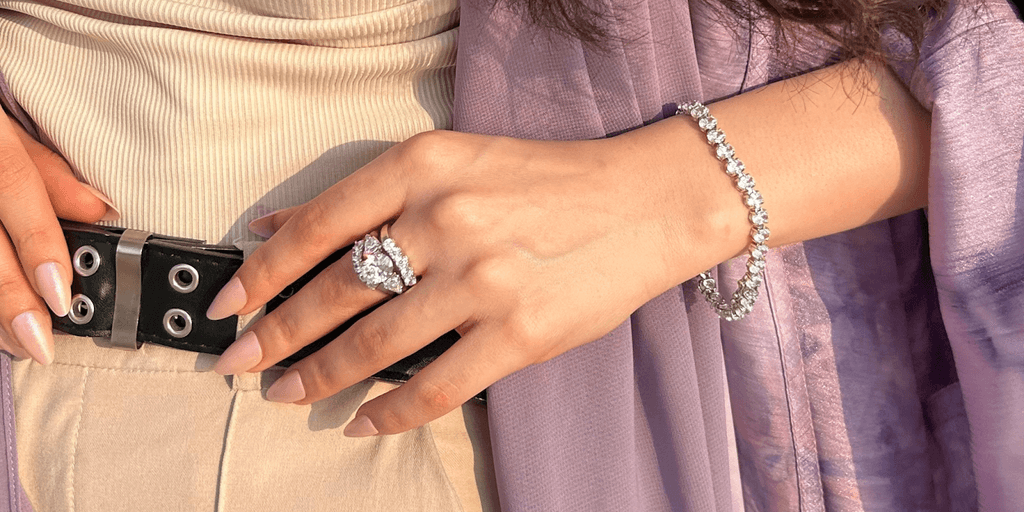 A close-up of a hand with a diamond ring and bracelet, wearing a beige top and black belt, with soft pink nails.