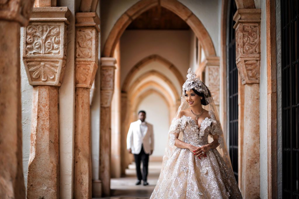 Bride in ornate gown smiles as groom approaches in arched, stone hallway.
