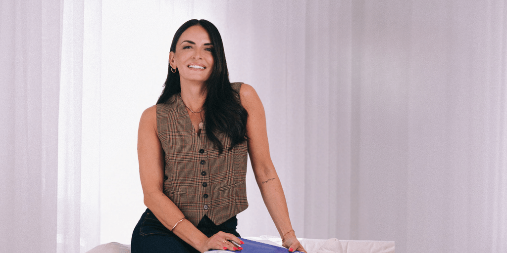 Smiling woman wearing a plaid vest and denim jeans, sitting on a couch with a white background, posing confidently