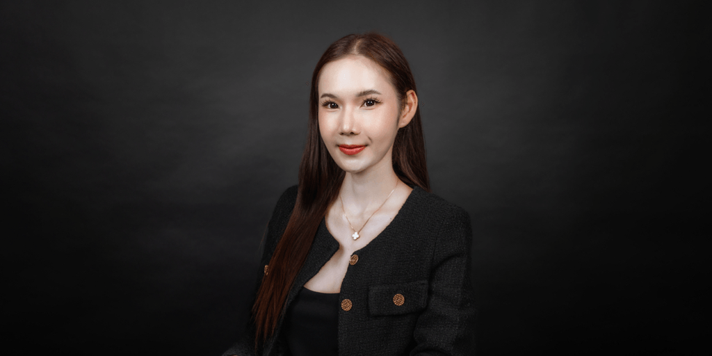 Portrait of a woman wearing a black blazer with gold buttons, a delicate necklace, and a confident smile, against a dark background.