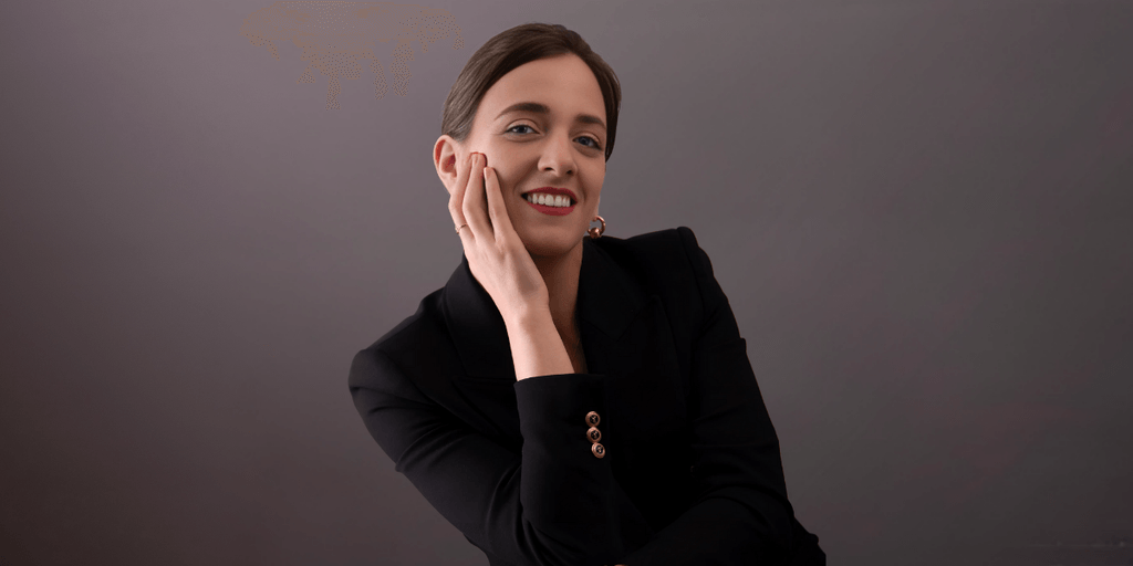 Professional woman in a black suit smiling while gently resting her hand on her face, against a neutral background.