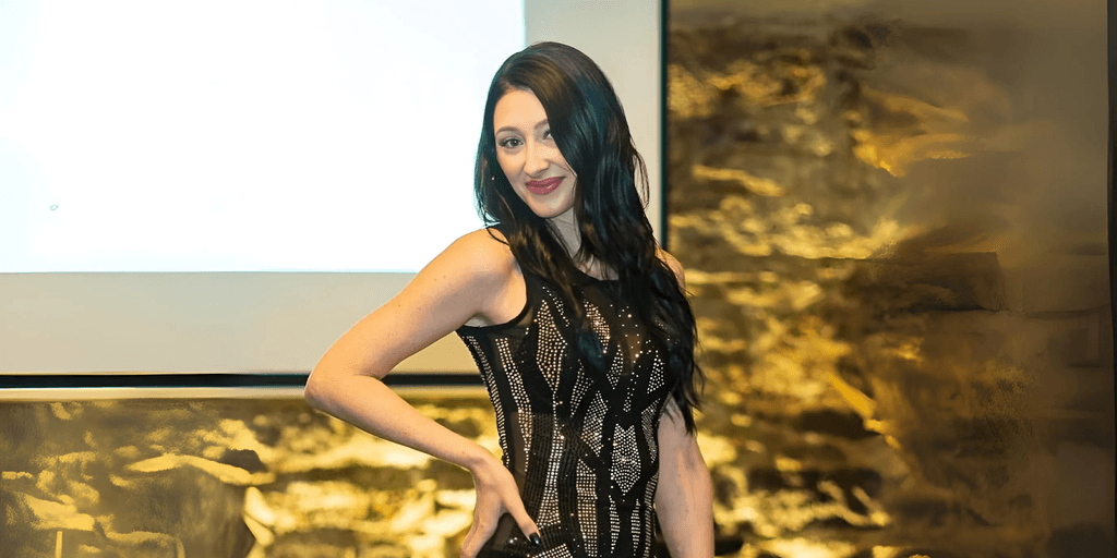 Woman in a black sequined dress posing confidently in front of a presentation screen and textured wall.