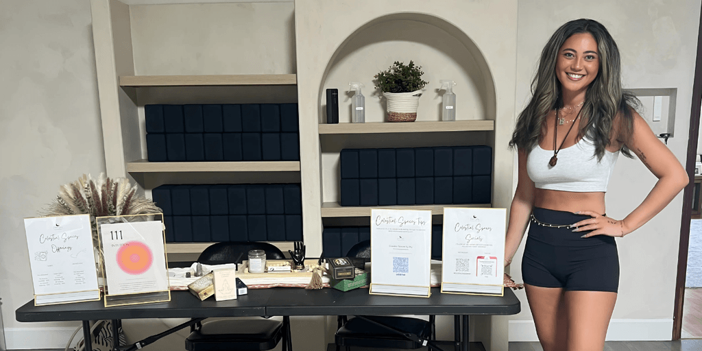 Smiling woman wearing a white crop top and black shorts, standing next to a display table with various crystal products and flyers for her business, with neatly arranged shelves and plants in the background.
