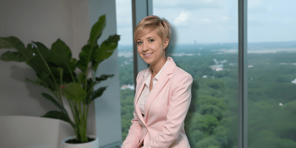 Professional woman with short blonde hair wearing a pink blazer and white blouse, sitting in front of a large window with a city view. She smiles confidently while seated next to a green plant in an office environment.