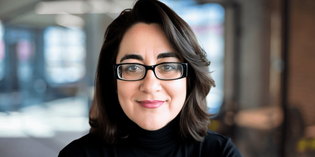 Portrait of a woman with brown hair and glasses, wearing a black turtleneck sweater, smiling confidently in a professional setting with blurred background.