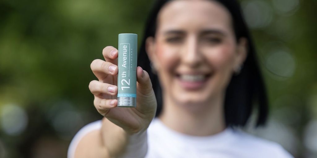 A smiling woman holding the 12th Avenue skincare HydraRetin balm stick in front of a blurred natural background, promoting its dual-action formula for hydration and skin care."
