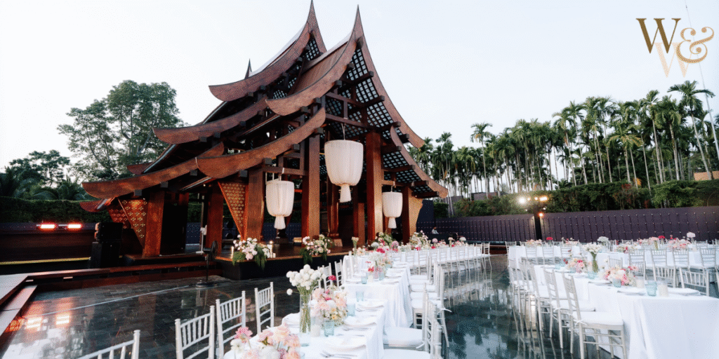 Outdoor wedding venue with a traditional wooden pavilion, long dining tables adorned with floral arrangements, and a scenic backdrop of palm trees under a clear sky.