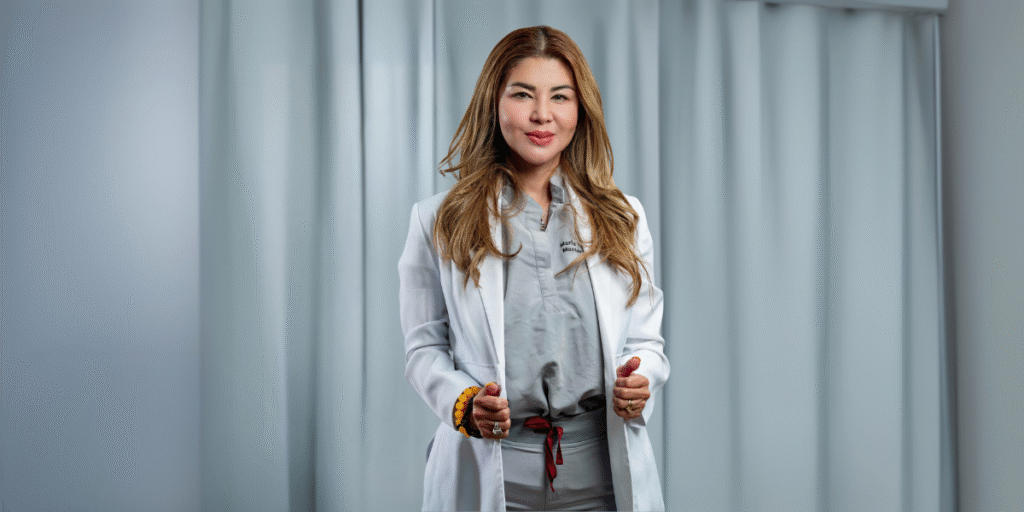 Professional woman in a white lab coat and grey medical scrubs standing confidently with a friendly expression. She is a skilled aesthetician, offering expertise in skincare treatments, with soft lighting and a neutral background.