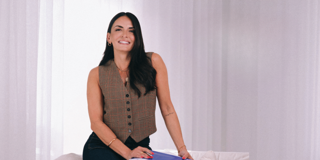 Smiling woman wearing a plaid vest and denim jeans, sitting on a couch with a white background, posing confidently