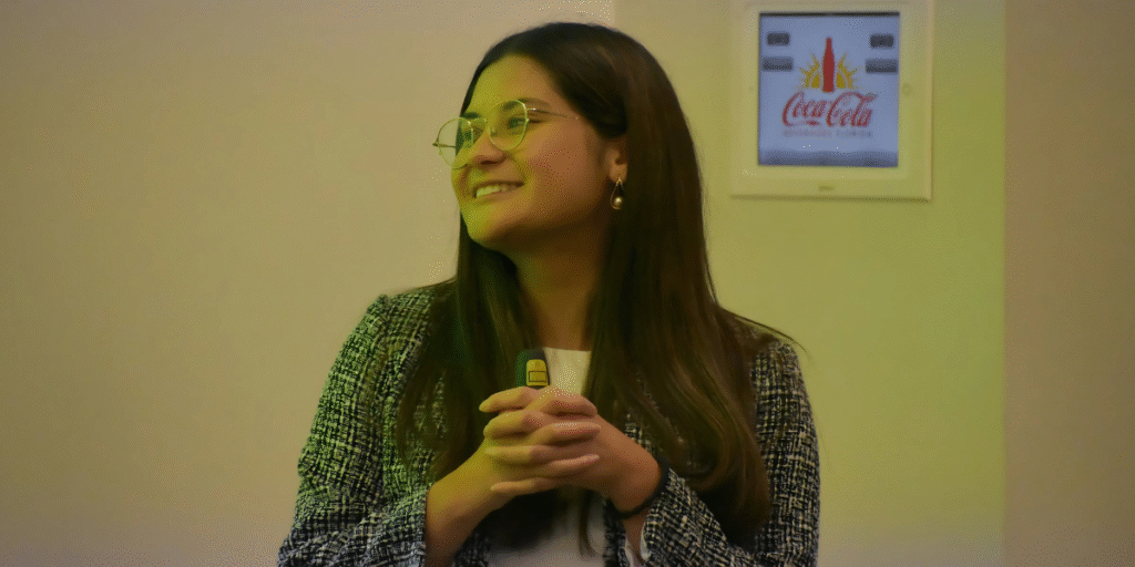 Young professional woman smiling and holding a remote control during a presentation, with a Coca-Cola advertisement visible in the background.