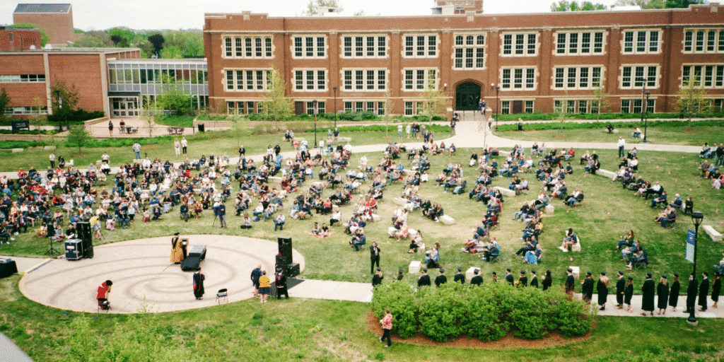 Outdoor graduation ceremony with a crowd of students and spectators gathered in front of a brick university building