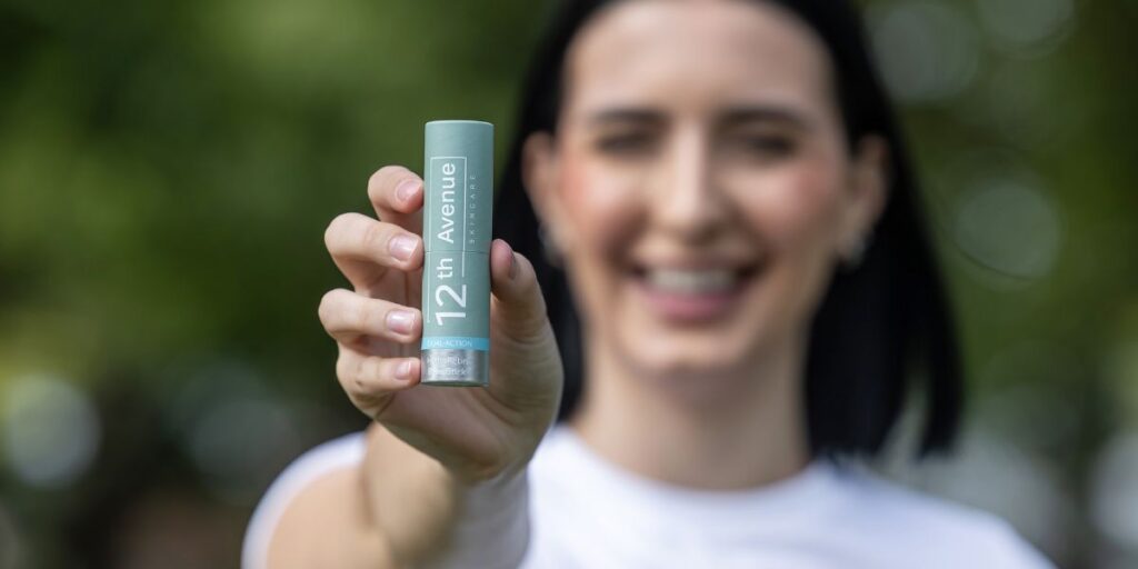 A smiling woman holding the 12th Avenue skincare HydraRetin balm stick in front of a blurred natural background, promoting its dual-action formula for hydration and skin care."