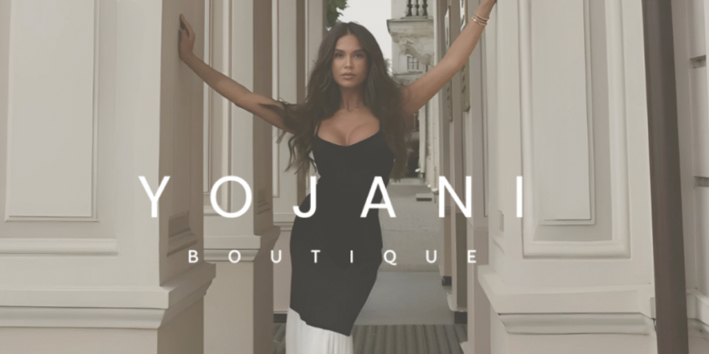 Fashion model posing confidently in a black dress, standing between elegant columns with the 'Yojani Boutique' logo displayed prominently, showcasing sophisticated and stylish clothing.