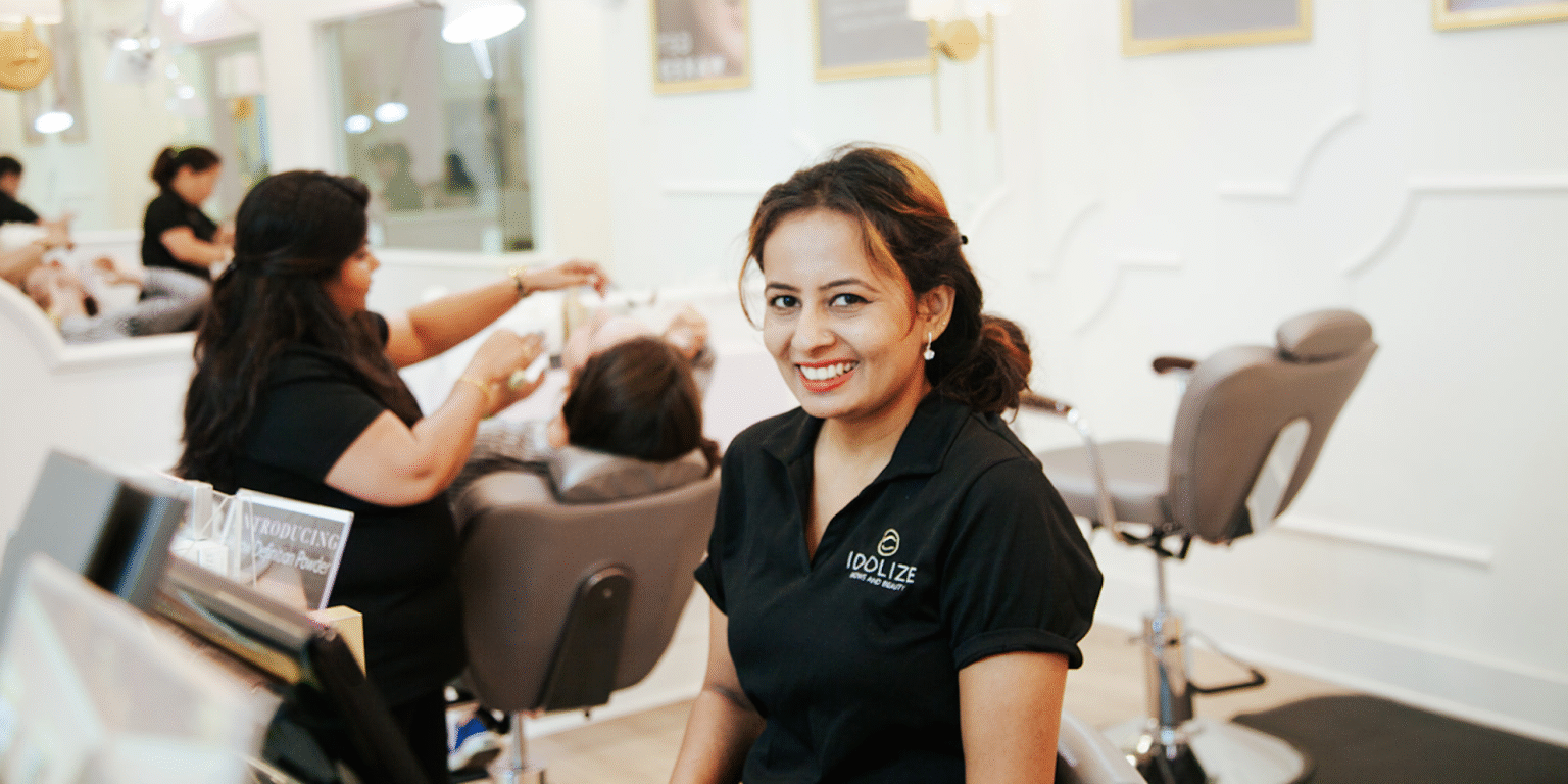 A woman in a black shirt with a logo on it is smiling while sitting at a beauty salon reception desk. In the background, other people are receiving beauty treatments.
