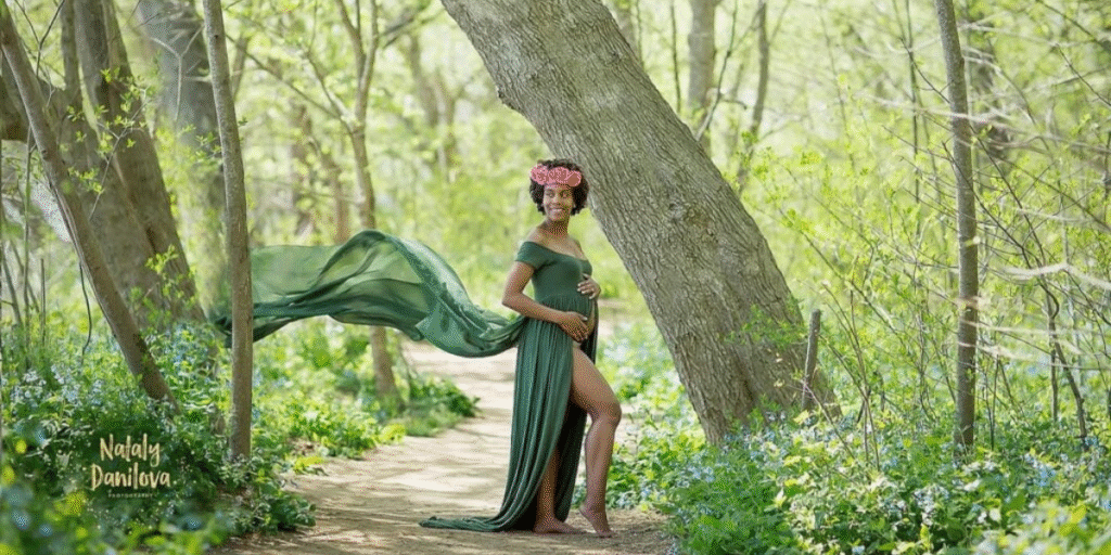 "Pregnant woman in a flowing green dress standing confidently in a nature-filled path with a flower crown, exuding elegance and grace during a maternity photo shoot."