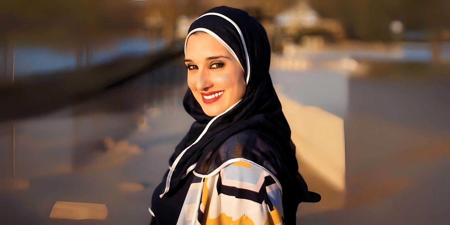 "Smiling woman in a colorful hijab and patterned coat, standing outdoors with a blurred background and sunny lighting."