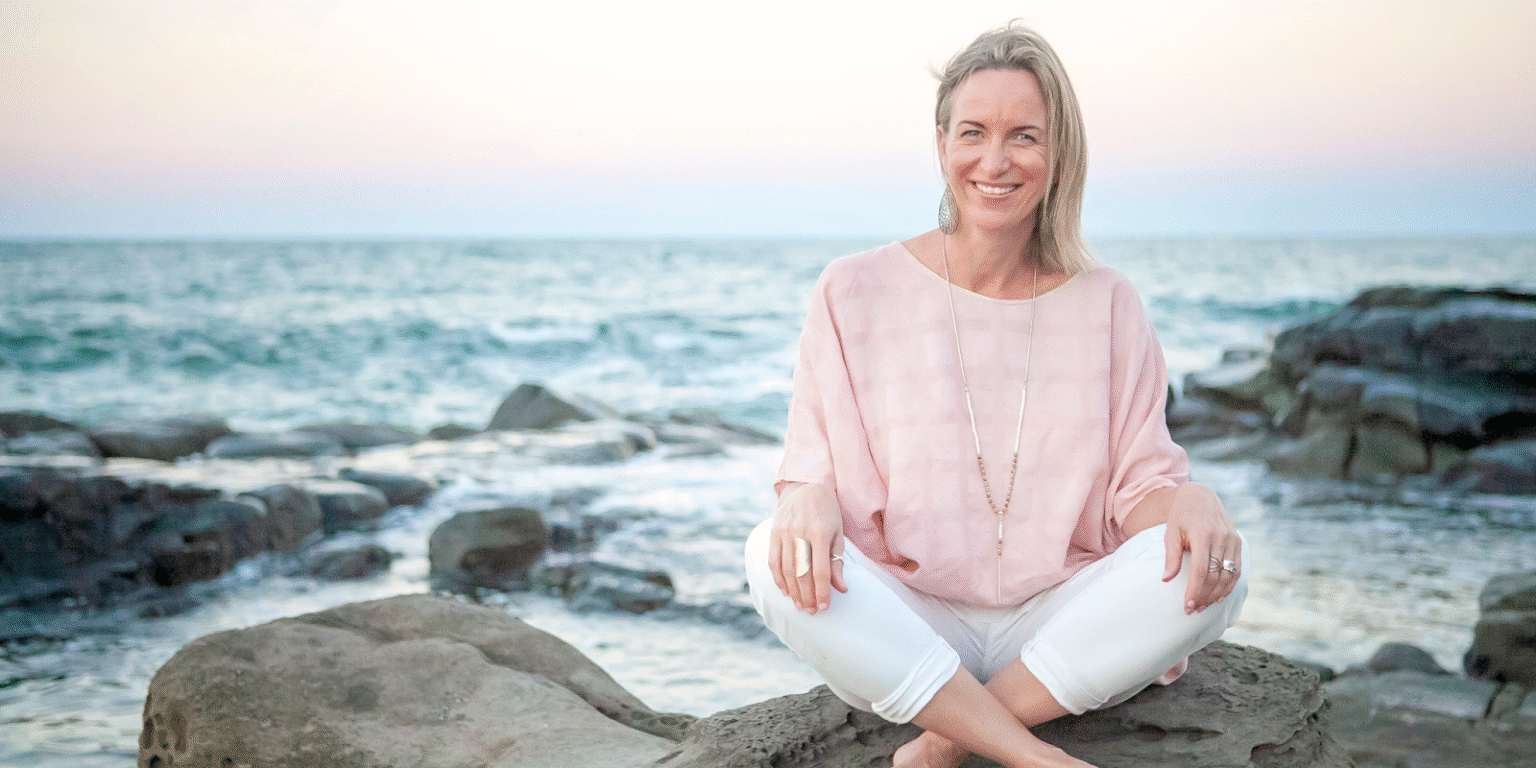 ChatGPT said: Woman in light pink blouse and white pants sitting cross-legged on rocks by the ocean at sunset.