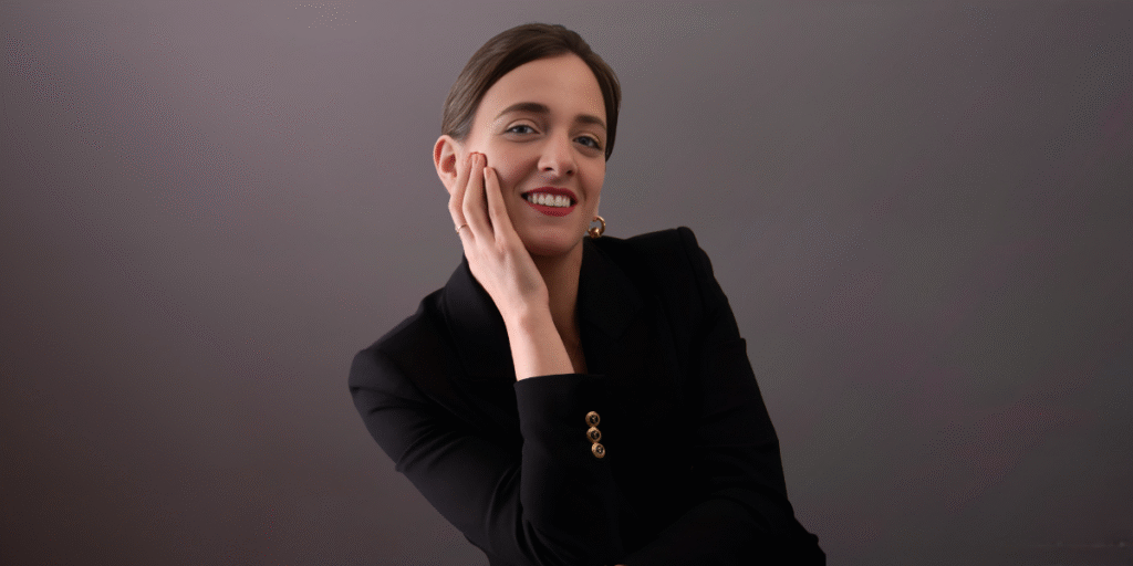 Professional woman in a black suit smiling while gently resting her hand on her face, against a neutral background.