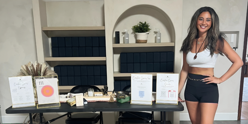 Smiling woman wearing a white crop top and black shorts, standing next to a display table with various crystal products and flyers for her business, with neatly arranged shelves and plants in the background.