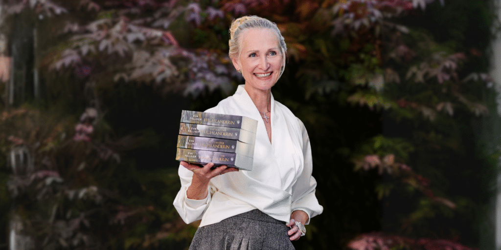 Eva Fellner smiling while holding a stack of The Highlanderess Saga books against a leafy background.