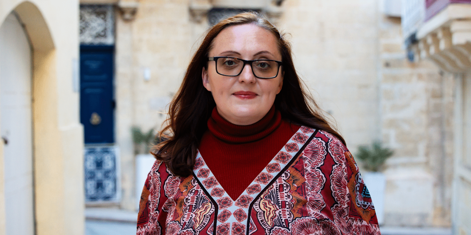 Woman dressed in a red turtleneck and patterned blouse, walking through an old city street with a traditional architectural backdrop.