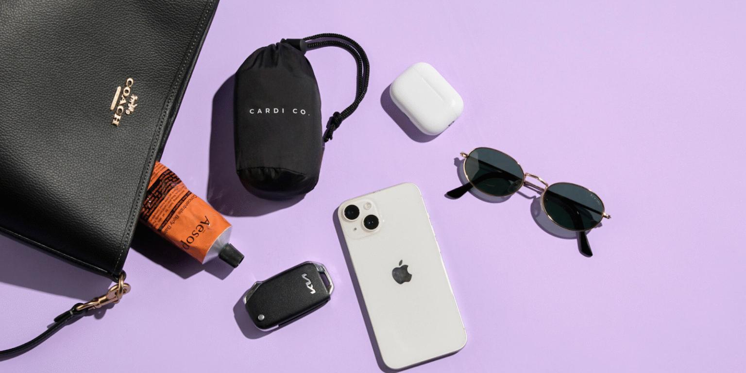 A flat lay of a black Coach handbag, a tube of Aesop hand cream, a "CARDI CO." branded bag, Apple AirPods, a phone, and sunglasses on a purple background.