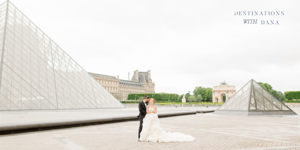 A couple in wedding attire embraces in front of the Louvre Pyramid in Paris, with the Eiffel Tower in the background.