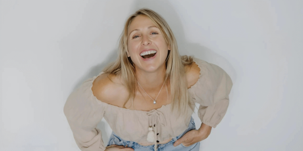 Libby Finlayson smiling with a joyful expression, wearing a beige off-shoulder blouse, and light blue jeans.