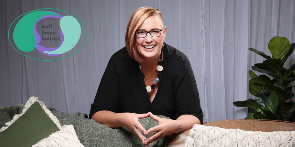 "Smiling woman in black shirt and colorful necklace, laughing while sitting on a couch surrounded by pillows, with the 'Well Being Holistic' logo in the corner."