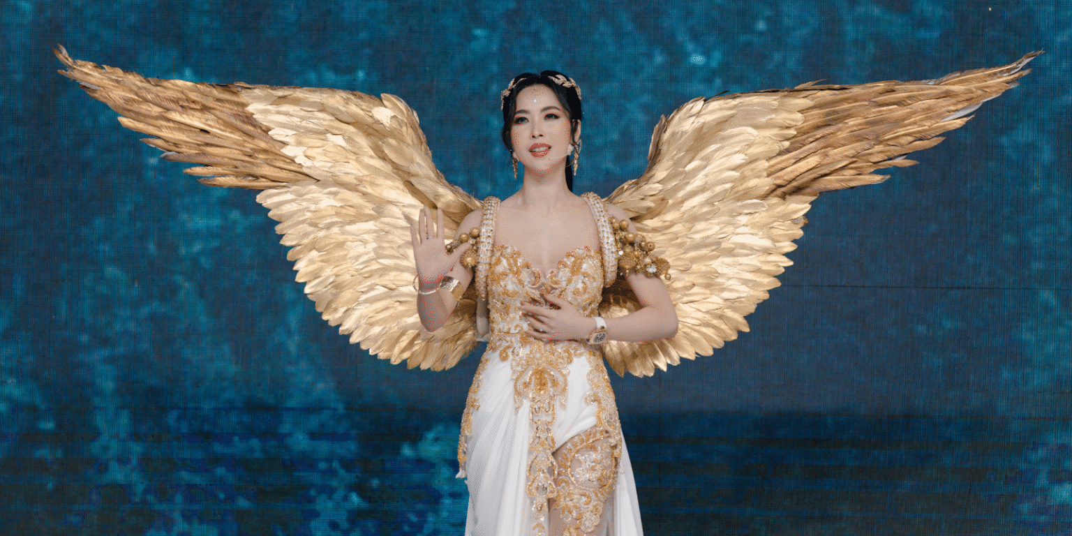 "Close-up of a model dressed in a glamorous angel-inspired costume with golden wings, giving a serene pose at a fashion show on a blue-lit stage."