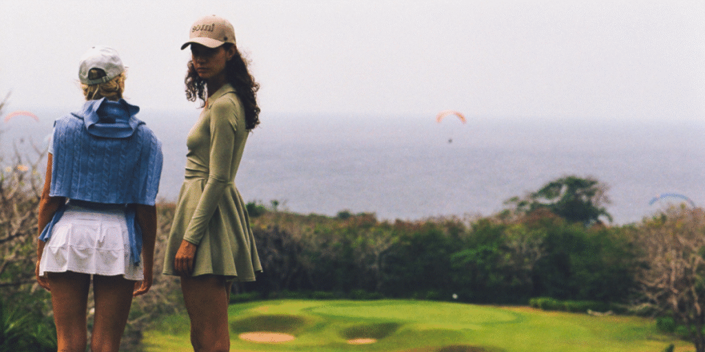 Two women in luxury headwear, overlooking a golf course with paragliders in the background, embodying elegant adventure.