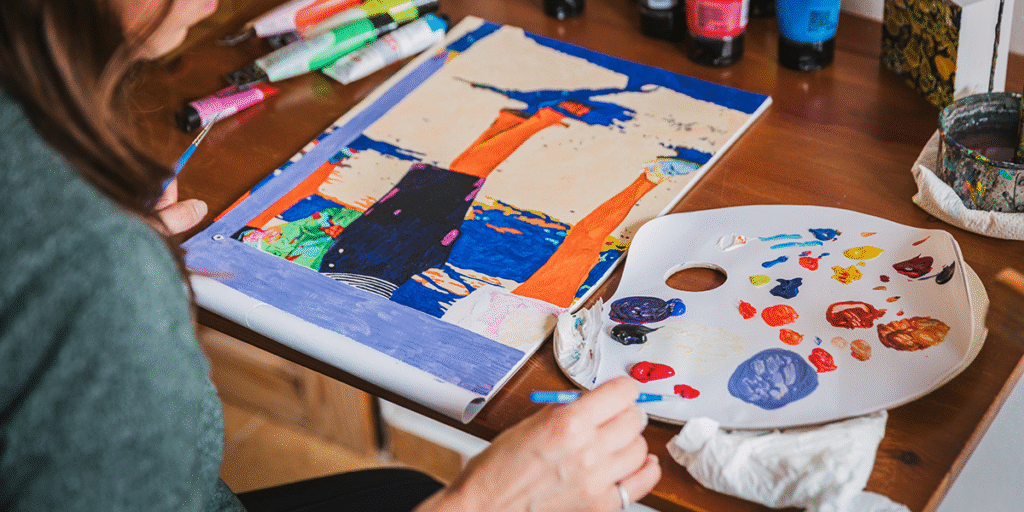 Artist painting a vibrant, colorful artwork at a desk with paint palette and supplies, reflecting creative expression.