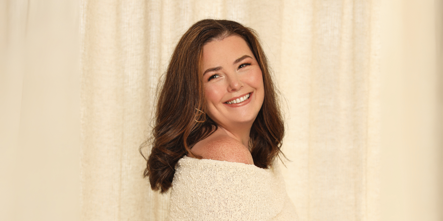 "Portrait of a woman smiling over her shoulder in a soft off-white sweater with wavy brown hair."