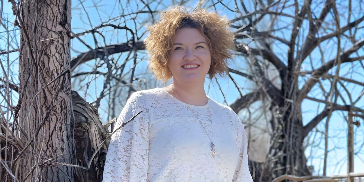 Danielle Dahl standing outside among bare trees, smiling, wearing a white top, symbolizing resilience and healing