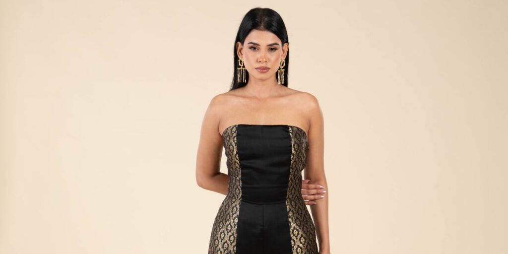 A South Asian woman models a black and gold strapless gown with intricate lace details and bold gold earrings.