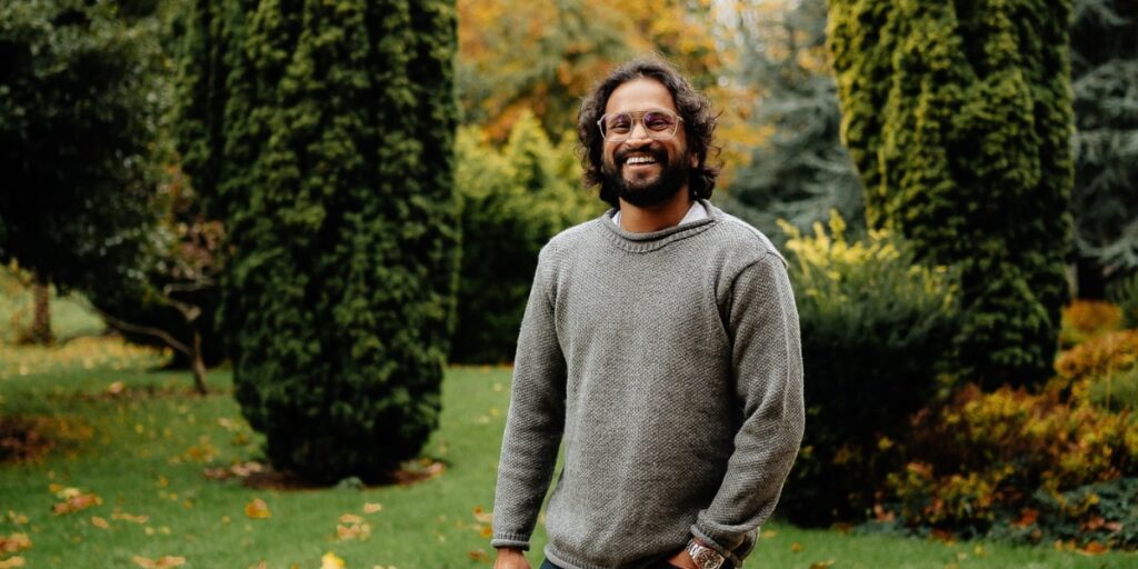 A smiling man with glasses stands outdoors amidst green trees, reflecting a warm, approachable presence that connects with nature.