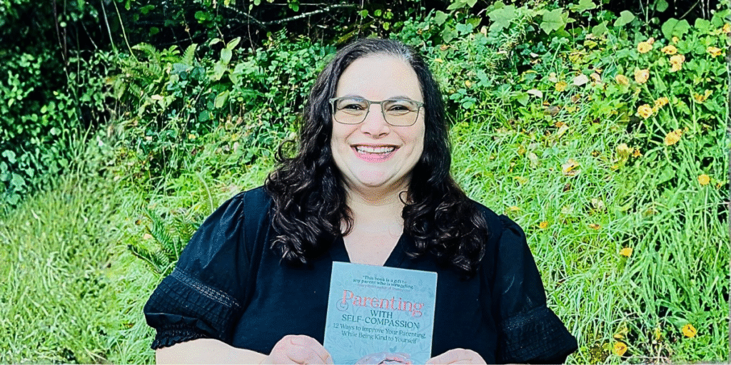Dr. Jen Ferris smiling while holding her book 'Parenting with Self-Compassion' in front of a green, flower-filled background, promoting her approach to compassionate parenting