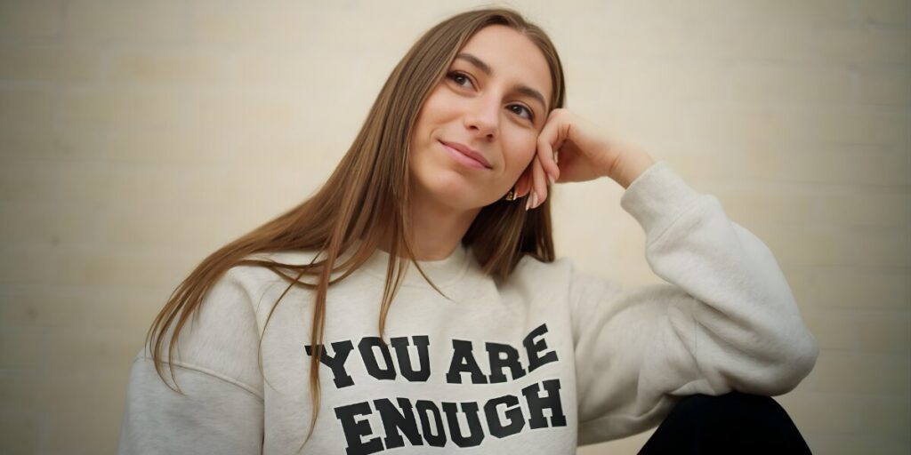Young woman with long hair, wearing a "You Are Enough" sweatshirt, sits pensively with a gentle smile. The background is a soft, neutral color.